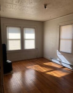 Empty sunlit room with wooden floors and two windows.