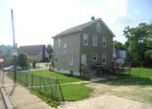 Two-story beige house with a grassy front yard and a chain-link fence.