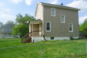A beige two-story house with a fenced yard and green grass.
